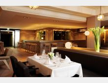 Elegantes Restaurant mit modernem Interieur, das eine gemütliche Bar mit hölzernen Barhockern und eine gedeckte Tafel mit weißen Servietten und Gläsern zeigt. Dekorative Elemente wie Tulpen in Vasen und warmes Licht schaffen eine einladende Atmosphäre.