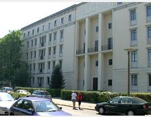 Wohngebäude im Stil des sozialistischen Klassizismus mit vier Etagen, symmetrischen Fenstern und Eingangsbereichen mit Säulen. Im Vordergrund geparkte Autos, ein Gehweg und zwei Personen, die die Straße entlanggehen.