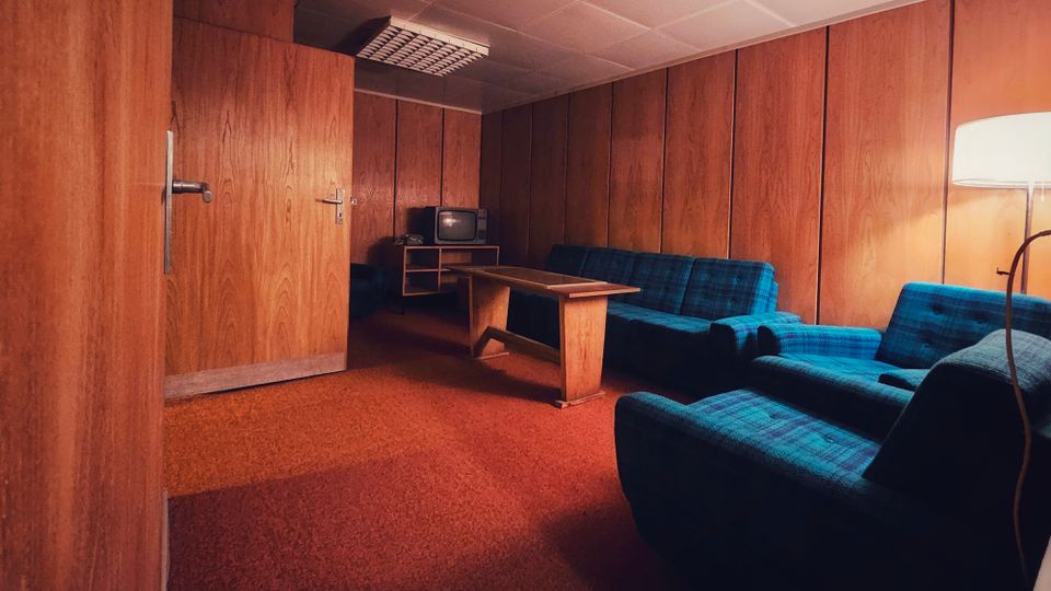 Holzvertäfelter Aufenthaltsraum mit rotem Teppichboden, abgehängter Decke mit Rasterleuchten und einem langen Holztisch in der Mitte. An den Wänden stehen blau karierte Sofas und Sessel, daneben ein kleiner TV auf einem Regal sowie eine Stehlampe.