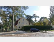 Zweistöckiges Einfamilienhaus mit gelber Fassade und rotem Satteldach, umgeben von hohen Bäumen und einer gepflegten Hecke; im Vordergrund ein geparkter blauer Kleinwagen auf einem gepflasterten Parkplatz.