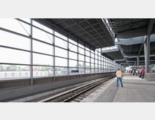 Ein moderner Bahnhof mit überdachten Bahnsteigen und weitläufigen Glaswänden, die Tageslicht einlassen. Auf dem Schild ist die Station „Berlin Südkreuz“ zu erkennen, und Menschen warten entlang der Gleise.