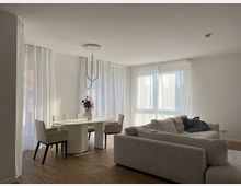 Helles, modernes Wohnzimmer mit großen Fenstern, durch die Tageslicht einfällt, weißen lichtdurchlässigen Vorhängen, einem runden Esstisch mit vier Stühlen und einer Couch in beigefarbenen Tönen. Der Boden besteht aus Holz und im Hintergrund steht eine Vase mit Blumen auf dem Tisch.