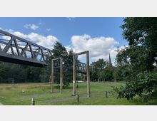 Eine Grünfläche mit vereinzelten Bäumen, auf der eine Stahlträgerbrücke verläuft. Im Vordergrund stehen mehrere Holzrahmen, im Hintergrund ist die Spitze eines Kirchturms zu sehen.