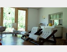 Wohnzimmer mit einer offenen, hellen Gestaltung, großen Fenstern mit Holzrahmen, die einen Blick ins Grüne bieten. Im Raum stehen zwei moderne, gepolsterte Sessel mit Holzkonstruktion, ein Couchtisch aus Holz, und im Hintergrund ist ein Sideboard mit Dekoration, darunter ein gelbes Sparschwein, sichtbar.