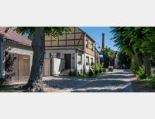 Fachwerkhaus mit heller Fassade und Blumen auf den Fensterbänken, an einer mit Bäumen gesäumten Kopfsteinpflasterstraße. Im Hintergrund ist ein hoher Schornstein sowie ein Gebäude mit einem Geschäftsschild erkennbar.