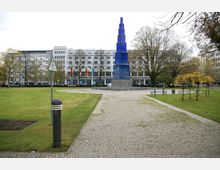 Ein Stadtpark mit gepflasterten Wegen, Rasenflächen und Bäumen. In der Mitte steht eine hohe blaue Säulenskulptur auf einem Sockel. Im Hintergrund sind moderne Gebäude und Fahnen zu sehen.