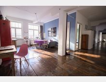 Weitläufiger Wohnraum mit Holzdielenboden und großen Fenstern, die viel Tageslicht hereinlassen. Der Raum ist teils in Blau gestrichen, mit einem Esstisch, einer roten Kühlschrankfront und einer Sitzecke ausgestattet, sowie einem offenen Übergang zu weiteren Zimmern.