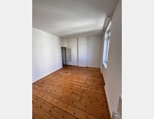 Helles, leeres Zimmer mit Holzdielenboden, weißen Wänden und einer Tür mit Glaseinsatz. Rechts befindet sich ein großes Fenster, das Tageslicht in den Raum lässt.