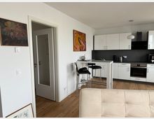 Moderne Küchen- und Wohnraumkombination mit weißer Einbauküche, schwarzen Arbeitsplatten und Holzboden. Im Vordergrund ein heller Polstersessel, im Hintergrund ein Tresen mit schwarzen Barhockern; an den Wänden hängen Gemälde.