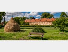 Eine ländliche Szene mit einer Holzbank im Vordergrund, einem großen Heuhaufen links und einem Fachwerkgebäude mit rotem Ziegeldach im Hintergrund, umgeben von grünen Wiesen, Bäumen und einem klaren Himmel.
