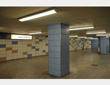 U-Bahnstation mit gefliesten Wänden in Weiß, Blau und Orange, einem beleuchteten Schild mit der Aufschrift „Rathaus Steglitz U9“ und mehreren quadratischen Säulen, die mit blauen Fliesen verkleidet sind. Die Decke ist flach mit langen Leuchtstofflampen, und der Boden besteht aus grauen Fliesen.