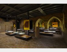 Innenansicht eines historischen Gewölberaums mit mehreren Spitzbogenarkaden und Backsteinwänden. Der Raum ist mit modernen Ausstellungsvitrinen und beleuchteten Exponaten ausgestattet, ergänzt durch gelb hinterlegte Wandbilder mit schwarzen Symbolen und Figuren.