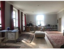 Großes Büro oder Arbeitszimmer mit Holzdielenboden und mehreren Teppichen, ausgestattet mit Schreibtischen, Regalen voller Aktenordner und großen Fenstern mit roten Vorhängen. Tageslicht fällt durch die Fenster und erhellt den Raum.