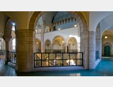 Offener Innenraum eines historischen Gebäudes mit Rundbögen, Säulen aus Stein und schmiedeeisernen Geländern. Die Wände sind in hellen Farben gehalten, und die Decke ist mit bläulichen Mustern dekoriert.