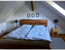 Schlafzimmer unter dem Dach mit schrägen weißen Wänden, einem Holzbett mit natürlichem Kopfteil, Bettwäsche mit floralem Muster, zwei kleinen Holznachttischen mit schwarzen Lampen und einem Teppich auf Holzfußboden.
