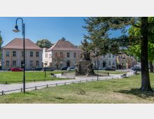 Zentraler Platz mit Rasenflächen und gepflasterten Wegen, in der Mitte ein barockes Denkmal mit Skulpturen, umgeben von Bäumen und historischen Gebäuden mit roten Ziegeldächern. Parkbänke und Straßenlaternen säumen die Wege; vereinzelt parken Autos entlang der Straße.