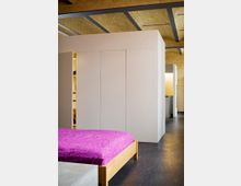 Modernes Zimmer in einem Loft mit hellem Backsteingewölbe und sichtbaren Stahlträgern an der Decke. Im Vordergrund ein Bett mit pinker Tagesdecke, im Hintergrund weiße, minimalistische Einbauten und dunkler Holzfußboden.