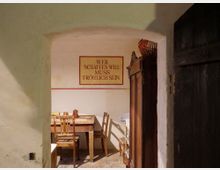 Blick in einen rustikalen Raum mit weiß verputzten Wänden, einem Holztisch und mehreren Stühlen. An der Wand hängt ein Schild mit der Aufschrift „Wer schaffen will muss fröhlich sein“, daneben ein Holzschrank und Details einer geöffneten Holztür.