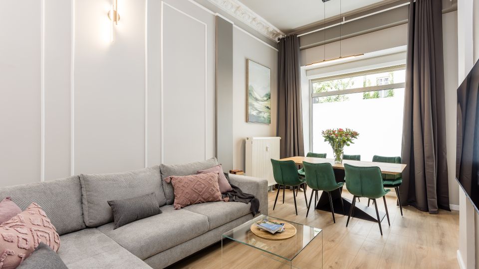 Modernes Wohn- und Esszimmer mit grauem Ecksofa, gläsernem Couchtisch und hellem Holzboden. Im Hintergrund steht ein Esstisch mit grünen Stühlen vor einem großen Fenster mit dunklen Vorhängen; Stuckleisten und Wandpaneele prägen den Raum.