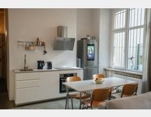 Moderne Küche mit weißen Schränken, eingebautem Herd und Dunstabzugshaube aus Edelstahl. Ein großer Kühlschrank mit integriertem Bildschirm steht neben einer Fensterfront, die Licht in den Raum lässt; im Vordergrund befindet sich ein Esstisch mit Holzstühlen.