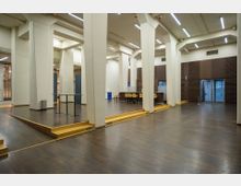 Innenraum eines modernen Veranstaltungsraums mit hohen weißen Säulen, dunklem Holzfußboden und abgetrennten Bereichen durch kleine Podeste. Im Hintergrund befinden sich eine Theke mit gelben Barstühlen und Wandelemente aus Holzverkleidung.