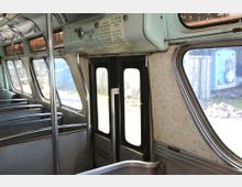 Innenansicht eines alten Busses, mit Metall-Sitzrahmen, großen Fenstern und einer zweiflügeligen Tür im Hintergrund. Die Wandverkleidung ist mit einem grünlichen Muster gestaltet, während an der Decke Schilder mit Verhaltensregeln angebracht sind.