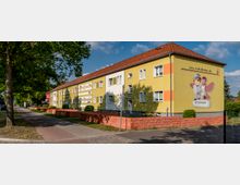 Gelbes Wohngebäude mit rotem Satteldach und weißen sowie orangefarbenen Wandakzenten, daneben ein Gehweg mit Bäumen und eine Straße. An der Fassade ist ein großes Werbemotiv mit Figuren und einer Internetadresse angebracht.