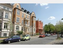 Straßenszene mit mehrstöckigen Wohnhäusern im Altbaustil, teils aus hellen Ziegeln und Putzfassaden, sowie einem auffälligen roten Gebäude im Hintergrund. Am Straßenrand stehen mehrere parkende Autos, und Bäume säumen den Gehweg unter einem klaren Himmel.