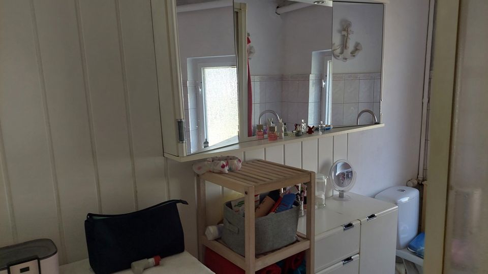 Kleines Badezimmer mit weiß vertäfelten Wänden, einem großen Spiegelschrank über einem Waschbecken und einem Milchglasfenster. Im Raum stehen ein Holzregal und ein weißer Unterschrank, rechts ist ein WC zu sehen.