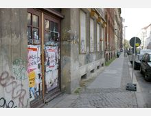Straßenszene mit einer Fassade eines älteren Gebäudes, das mit Graffiti und Plakaten bedeckt ist, und verschlossenen Fenstern mit heruntergelassenen Rollläden. Der Gehweg aus Pflastersteinen führt an parkenden Autos und weiteren Gebäuden entlang.