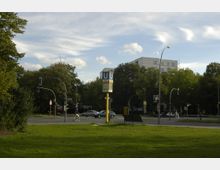 Eine Kreuzung in einer urbanen Umgebung mit einer U-Bahn-Säule, die das blaue "U"-Symbol und eine Uhr zeigt. Im Hintergrund sind Bäume, eine Straße mit Autos und ein modernes Gebäude mit weißen Fassaden sichtbar; im Vordergrund befindet sich eine Rasenfläche.