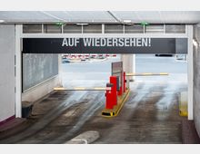 Ausfahrt einer Tiefgarage mit roten Schrankenanlagen und einem schwarzen Schild darüber mit der Aufschrift „AUF WIEDERSEHEN!“. Dahinter ist eine Straße mit Absperrungen und Bordsteinkanten sichtbar.