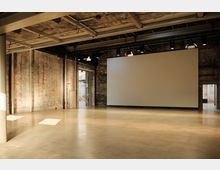 Industriehalle mit freiliegenden Backsteinwänden, einem großen leeren Raum mit glattem Betonboden und einer weißen Leinwand an der Wand. Das Gebäude verfügt über eine offene Stahlkonstruktion an der Decke und einen Türdurchgang, der zu einem weiteren Bereich führt.