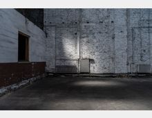 Ein leerer Raum mit einer grob verputzten weißen Ziegelwand, an der Heizkörper und Rohrleitungen angebracht sind. Der Boden ist aus dunklem Beton, und seitlich ist eine Wand mit einem kleinen, offenen Fenster zu sehen.