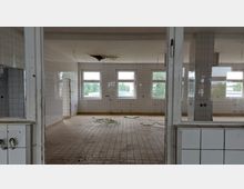 Ein leerer, verlassener Raum mit gefliesten Wänden und einem gefliesten Boden. Die Decke zeigt Schäden mit herausgebrochenen Stellen, und es hängen freiliegende Kabel herunter. Durch die Fenster ist grüne Vegetation im Außenbereich sichtbar.