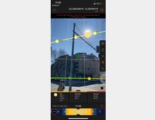 Screenshot einer App, die den Sonnenstand anzeigt. Im Hintergrund ist eine städtische Kreuzung mit einem Straßenmast, einer Verkehrsampel und einem mehrstöckigen Wohngebäude zu sehen. Der Himmel ist klar und die Sonne scheint hoch am Himmel.