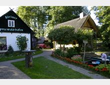 Ein idyllischer Hof mit einer weißen Fachwerkgebäudeaufschrift "Kleiner Spreewald Hafen", umgeben von Blumenschmuck, Sonnenblumen und einer Rasenfläche. Rechts im Bild steht ein rustikales Gebäude mit Strohdach, daneben eine Anlegestelle mit Holzbooten, Bäumen und Blumenbeeten.
