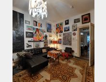 Wohnzimmer mit einer schwarzen Ledercouch, gemustertem Teppichboden und einer Vielzahl von Kunstwerken und Fotografien an den Wänden. Im Raum befinden sich ein kleiner Tisch mit gemustertem Tuch, Sessel und eine Stehlampe; im Hintergrund ist eine offene Tür zum angrenzenden Essbereich sichtbar.