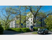 Wohnviertel mit mehrstöckigen, weißen Wohngebäuden, umgeben von Bäumen mit frischem Laub. Im Vordergrund sind ein Gehweg, parkende Autos und Straßenschilder zu sehen, darunter ein beleuchteter Briefkasten mit Werbung.