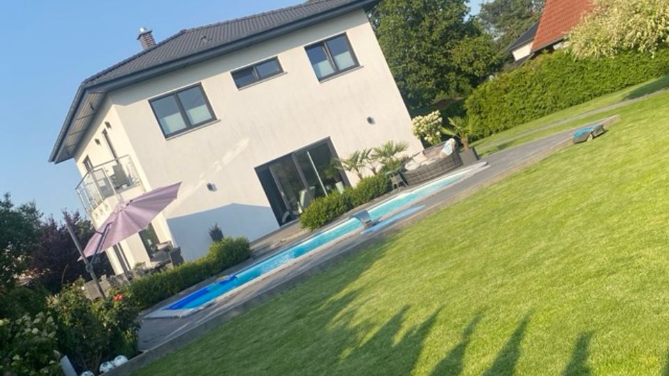 Modernes zweigeschossiges Wohnhaus mit grauem Satteldach und großen Fenstern, davor eine Terrasse mit Sonnenschirm und ein rechteckiger Pool. Im Vordergrund liegt ein großer, gepflegter Rasen, umgeben von Hecken und Bäumen in einem Wohngebiet.