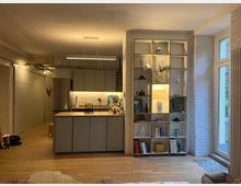 Offener, moderner Wohnraum mit integrierter Küche, bestehend aus grauen Küchenmöbeln, einer Kücheninsel und einer beleuchteten weißen Backsteinwand. Rechts befindet sich ein hohes Regal mit Büchern und Dekorationen, daneben eine Terrassentür mit Blick ins Freie.