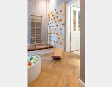 Ein heller Innenraum mit hölzernem Parkettboden, ausgestattet mit einer Kletterwand, einer Sprossenwand mit Schaukeln, einer Wippe aus Holz und einem Ballbecken. Rechts führt eine geöffnete Tür in einen weiteren Raum mit farbigem Wanddesign.