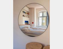 Ein modernes Schlafzimmer, reflektiert in einem großen, runden Spiegel. Zu sehen sind ein Bett mit weißer Bettwäsche, ein offenes Regal mit Büchern an der Wand sowie ein geöffnetes Fenster, das Tageslicht hereinlässt; im Vordergrund sind runde, geflochtene Hocker sichtbar.