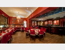 Ein festlich dekoriertes Restaurant mit roten Wänden, Holzboden und einem großen Kronleuchter. Tische sind mit weißen Tischdecken, roten Servietten und Stühlen mit roten Sitzhussen ausgestattet, daneben eine holzverkleidete Bar mit Regalen und Gläsern im Hintergrund.
