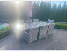 Terrasse mit großformatigen Steinplatten, einem rechteckigen Glastisch und sechs Korbstühlen; in der Mitte steht ein geschlossener Sonnenschirm. Im Hintergrund begrenzen eine niedrige Hecke und hohe dichte Nadelbäume den Gartenbereich.