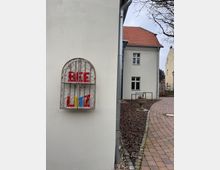 Eine Ecke eines Gebäudes mit heller Putzfassade, an der ein metallisch verziertes Dekoelement mit bunten Buchstaben angebracht ist. Rechts führt ein gepflasterter Weg entlang eines kleinen bepflanzten Streifens, und im Hintergrund sind weitere Gebäude mit roten Ziegeldächern sichtbar.