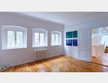 Heller Raum mit Holzdielenboden, drei großen Fenstern mit weißen Sprossenrahmen und einem Heizkörper darunter. An der Wand hängen vier blaue Bilder, und rechts führt eine offene Tür zu einem Flur mit einer weißen Holztreppe.