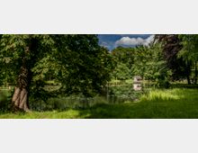 Parklandschaft mit einem kleinen See, umgeben von Bäumen. Auf dem See schwimmen Seerosen, und am Ufer steht ein kleines Holzhaus mit spitzem Dach.