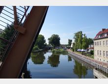 Blick auf einen Kanal mit ruhigem Wasser, gesäumt von Bäumen und einer Uferpromenade mit Geländern. Rechts im Bild stehen Gebäude und ein Café mit Sonnenschirmen, links ragt ein Teil einer metallischen Brückenstruktur ins Bild.
