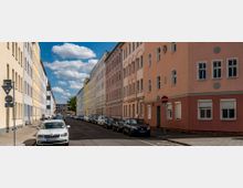 Straßenszene mit mehreren mehrstöckigen Wohnhäusern in Pastellfarben, die zu beiden Seiten der Straße stehen. Auf der Straße sind parkende Autos und am Rand sind Gehwege mit vereinzelten Verkehrsschildern erkennbar, im Hintergrund ein blauer Himmel mit Wolken.